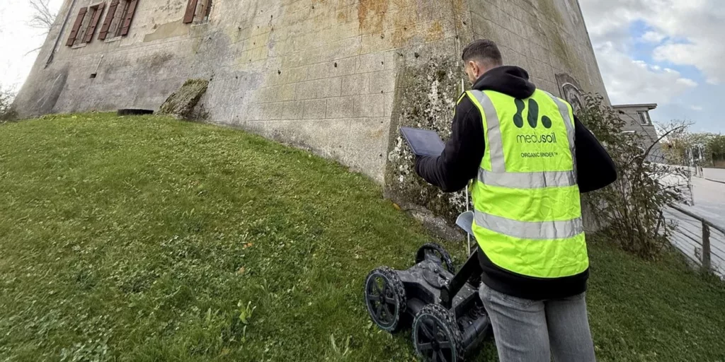 Medusoil engineer performing non-destructive ground monitoring with a wheeled geophysical scanner and tablet beside a historic building.